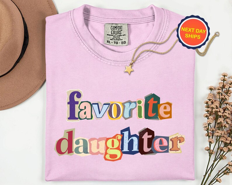 May include: A pale pink t-shirt featuring the words "favorite daughter" in a colourful, cut-out style. A gold necklace with a star pendant is on the shirt. A "Next Day Ships" sticker is visible.
