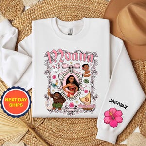 Puede incluir: Sudadera blanca con la palabra "Moana" en rosa, enmarcada por ilustraciones de personajes. La manga muestra el nombre "Jasmine" y una flor de hibisco rosa. Una pegatina de "Next Day Ships" es visible.