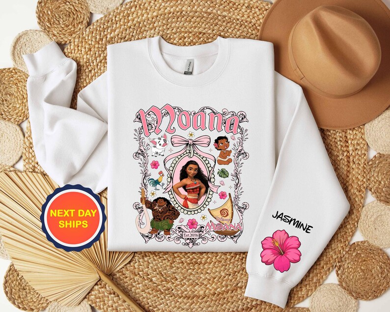 Puede incluir: Sudadera blanca con la palabra "Moana" en rosa, enmarcada por ilustraciones de personajes. La manga muestra el nombre "Jasmine" y una flor de hibisco rosa. Una pegatina de "Next Day Ships" es visible.