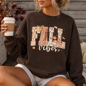 May include: A dark brown crewneck sweatshirt featuring a graphic print with the text "FALL vibes". The letters are filled with a pattern of autumn leaves, flowers, and pumpkins in orange, tan, and cream tones, accented by small bows and stars.