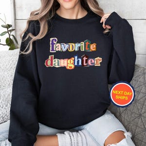 Puede incluir: Sudadera negra con las palabras "favorite daughter" en letras coloridas de estilo recortado. La sudadera tiene cuello redondo y mangas largas. Es visible una pegatina redonda con el texto "NEXT DAY SHIPS".