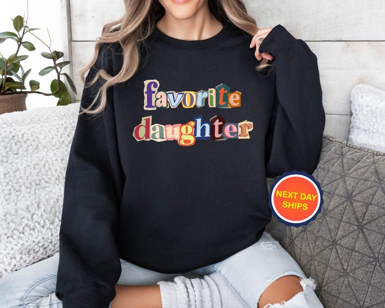 Puede incluir: Sudadera negra con las palabras "favorite daughter" en letras coloridas de estilo recortado. La sudadera tiene cuello redondo y mangas largas. Es visible una pegatina redonda con el texto "NEXT DAY SHIPS".