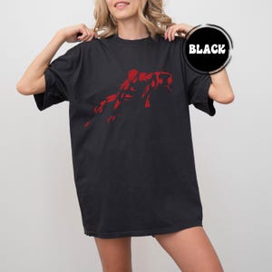 May include: A black short-sleeved t-shirt featuring a red graphic print of a crouching figure in the centre. The shirt has a crew neckline and a loose fit. A circular black badge with the white text BLACK is positioned in the upper right corner.