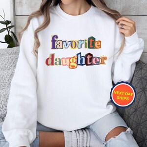 Puede incluir: Sudadera blanca con la frase "favorite daughter" en texto colorido y superpuesto, estilo recorte de revista. Una pegatina redonda con "NEXT DAY SHIPS" es visible en la parte inferior derecha.