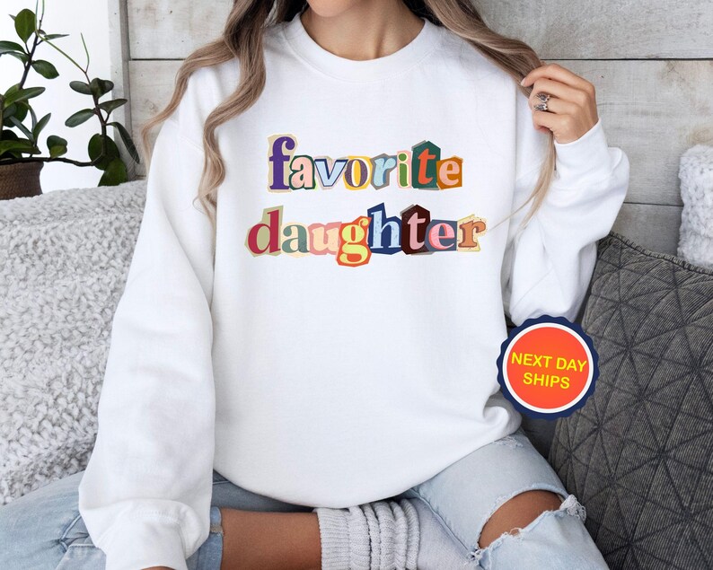 Puede incluir: Sudadera blanca con la frase "favorite daughter" en texto colorido y superpuesto, estilo recorte de revista. Una pegatina redonda con "NEXT DAY SHIPS" es visible en la parte inferior derecha.