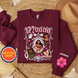 Puede incluir: Sudadera burdeos con un gr&aacute;fico del personaje Moana, con el nombre "Moana" en escritura rosa. La manga tiene el nombre "Jasmine" y una flor de hibisco rosa. La sudadera tiene cuello redondo.