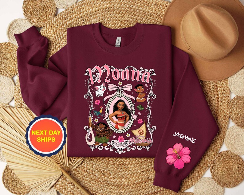 Puede incluir: Sudadera burdeos con un gr&aacute;fico del personaje Moana, con el nombre "Moana" en escritura rosa. La manga tiene el nombre "Jasmine" y una flor de hibisco rosa. La sudadera tiene cuello redondo.