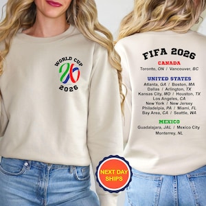 May include: Beige sweatshirt with the "World Cup 2026" logo on the front. The back features "FIFA 2026" and the host cities: Toronto, Atlanta, Dallas, Kansas City, Los Angeles, New York, Philadelphia, Bay Area, Guadalajara, and Monterrey.