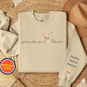 May include: A beige sweatshirt with the words "grandma bear" in script, featuring a cartoon bear with a honey pot and bees. The sleeve has the names Sophia, James, and Amelia.