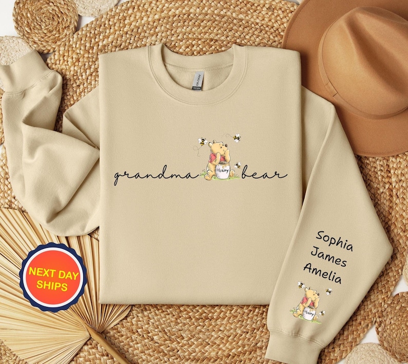 May include: A beige sweatshirt with the words "grandma bear" in script, featuring a cartoon bear with a honey pot and bees. The sleeve has the names Sophia, James, and Amelia.