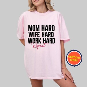 May include: A light pink t-shirt with the text "MOM HARD WIFE HARD WORK HARD Repeat" in black and pink. The shirt has short sleeves and a crew neck. A round sticker with the text "NEXT DAY SHIPS" is on the bottom right.