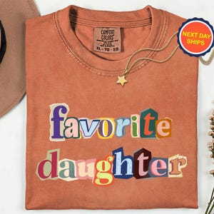 May include: A terracotta-coloured t-shirt featuring the words "favorite daughter" in colourful, cut-out style letters. A gold star pendant necklace and a "Next Day Ships" sticker are also visible.