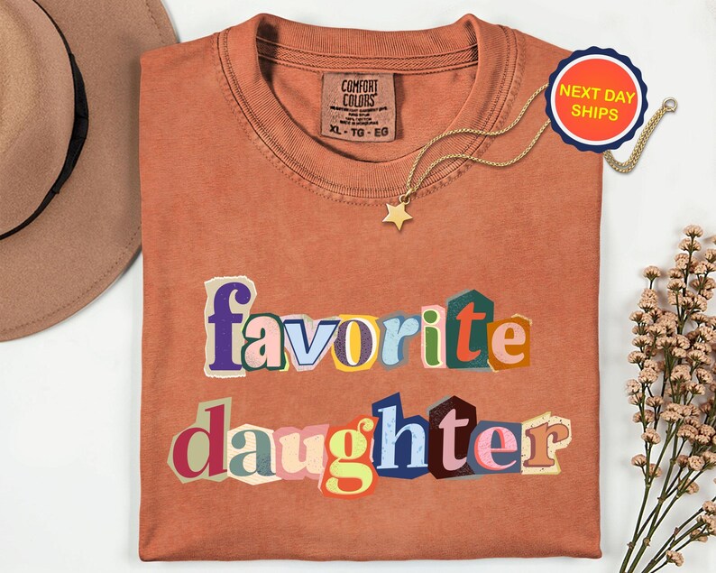 May include: A terracotta-coloured t-shirt featuring the words "favorite daughter" in colourful, cut-out style letters. A gold star pendant necklace and a "Next Day Ships" sticker are also visible.