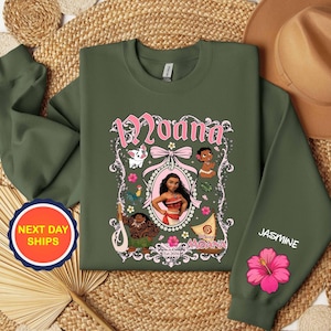 Puede incluir: Sudadera verde oliva con un gr&aacute;fico rosa y blanco del personaje Moana. El dise&ntilde;o incluye el nombre "Moana", ilustraciones de personajes y el texto "Est. 2016". La manga tiene el nombre "Jasmine" y una flor de hibisco rosa.