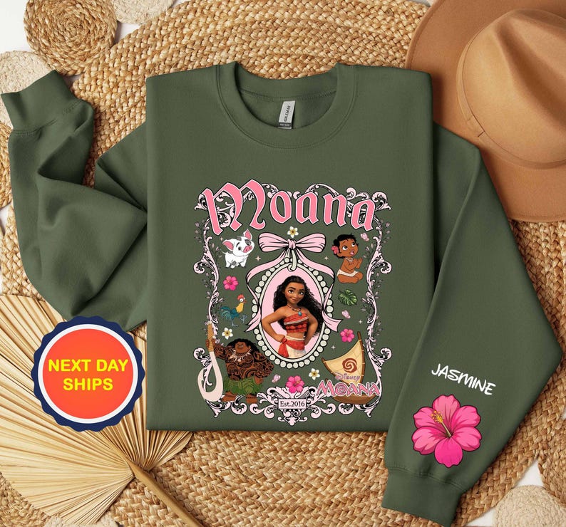 Puede incluir: Sudadera verde oliva con un gr&aacute;fico rosa y blanco del personaje Moana. El dise&ntilde;o incluye el nombre "Moana", ilustraciones de personajes y el texto "Est. 2016". La manga tiene el nombre "Jasmine" y una flor de hibisco rosa.