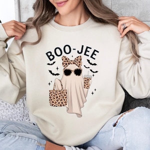 May include: A cream-colored crewneck sweatshirt featuring a graphic of a ghost wearing leopard print sunglasses and a matching bow. The ghost holds a leopard print tote bag and a matching cup, with the text BOO-JEE and black bat silhouettes.