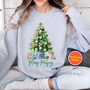 May include: A light gray sweatshirt featuring a Christmas tree decorated with pink bows and Mahjong tiles. The tree is set in a blue and white porcelain vase, with wrapped gifts at the base. The words "Merry Mahjong" are written in green script.
