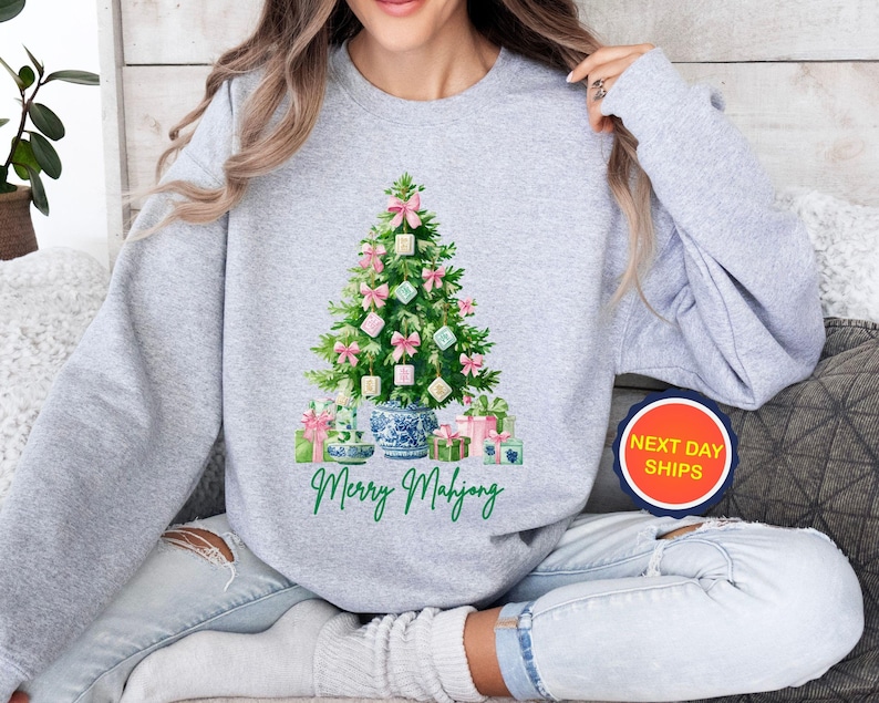 May include: A light gray sweatshirt featuring a Christmas tree decorated with pink bows and Mahjong tiles. The tree is set in a blue and white porcelain vase, with wrapped gifts at the base. The words "Merry Mahjong" are written in green script.