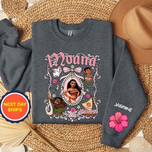 Puede incluir: Una sudadera gris oscuro con un gr&aacute;fico rosa y blanco del personaje Moana, otros personajes y el texto "Moana". La manga muestra el nombre "Jasmine" y una flor de hibisco rosa.