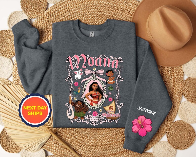 Puede incluir: Una sudadera gris oscuro con un gr&aacute;fico rosa y blanco del personaje Moana, otros personajes y el texto "Moana". La manga muestra el nombre "Jasmine" y una flor de hibisco rosa.