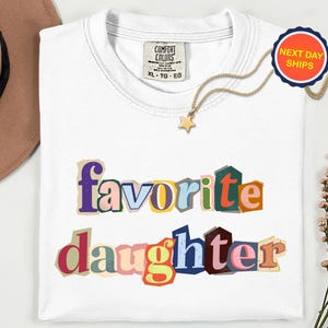 May include: A white t-shirt with the words "favorite daughter" in colourful, cut-out style letters. A gold necklace with a star pendant and a "Next Day Ships" sticker are also visible.
