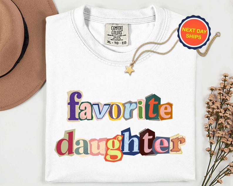 May include: A white t-shirt with the words "favorite daughter" in colourful, cut-out style letters. A gold necklace with a star pendant and a "Next Day Ships" sticker are also visible.