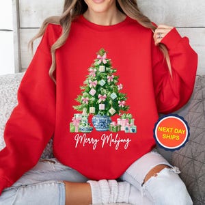 May include: A red sweatshirt featuring a Christmas tree made of Mahjong tiles, pink bows, and wrapped gifts. A blue and white vase is at the base of the tree. The words "Merry Mahjong" are written in cursive below the tree.