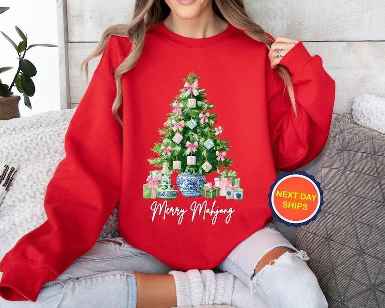 May include: A red sweatshirt featuring a Christmas tree made of Mahjong tiles, pink bows, and wrapped gifts. A blue and white vase is at the base of the tree. The words "Merry Mahjong" are written in cursive below the tree.