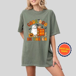 May include: Olive green t-shirt with a Halloween-themed graphic. The design features the text "CODE BOO IN THE ER" in orange and green, along with a ghost wearing a nurse's cap, a pumpkin, and bats. The shirt has a relaxed fit.