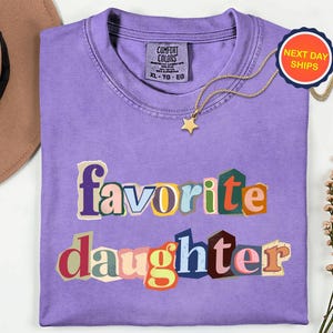 May include: A lilac Comfort Colors t-shirt with the words "favorite daughter" in a colourful, cut-out style. A gold star pendant necklace and a "Next Day Ships" sticker are also visible. A tan hat and flowers are in the background.