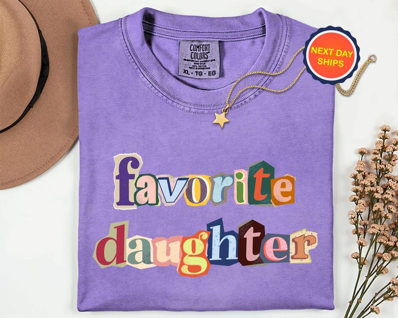 May include: A lilac Comfort Colors t-shirt with the words "favorite daughter" in a colourful, cut-out style. A gold star pendant necklace and a "Next Day Ships" sticker are also visible. A tan hat and flowers are in the background.