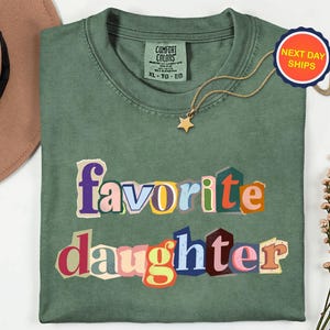 May include: A sage green t-shirt with the words "favorite daughter" in a colourful, cut-out style font. A gold star pendant necklace and a "Next Day Ships" sticker are also visible.