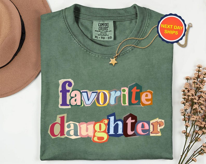 May include: A sage green t-shirt with the words "favorite daughter" in a colourful, cut-out style font. A gold star pendant necklace and a "Next Day Ships" sticker are also visible.