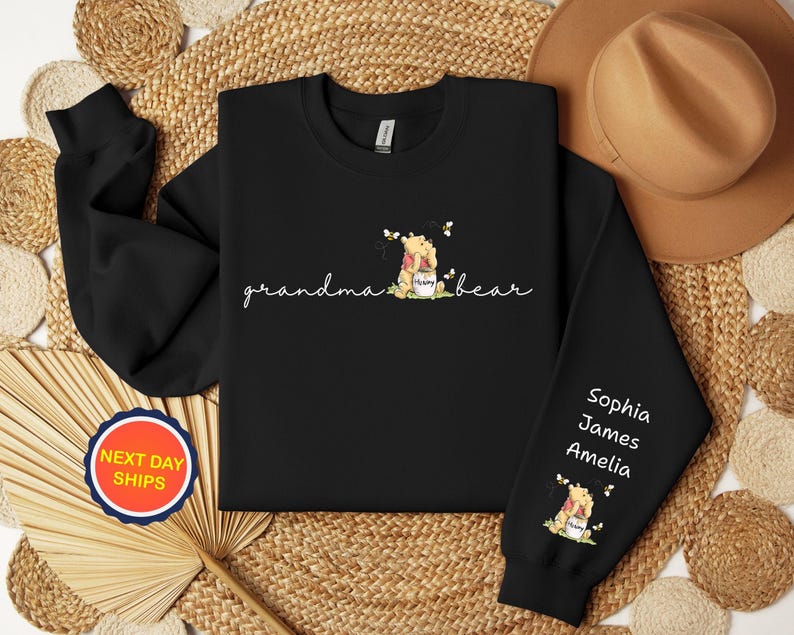 May include: Black sweatshirt with the words "grandma bear" in cursive, and a graphic of a bear holding a honey pot. The sleeve has the names Sophia, James, and Amelia. A round sticker says "NEXT DAY SHIPS".