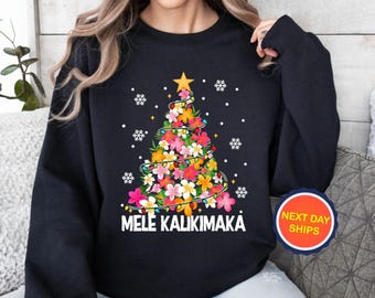 May include: Black sweatshirt featuring a Christmas tree design made of colorful flowers, a gold star, and string lights. The words "MELE KALIKIMAKA" are printed in white. White snowflakes are scattered around the tree.