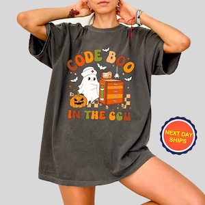 May include: Dark gray t-shirt with a Halloween-themed graphic. The design features a ghost wearing a nurse's hat, a pumpkin, and the text "CODE BOO IN THE CCU" in orange and brown. The shirt has a vintage, worn look.