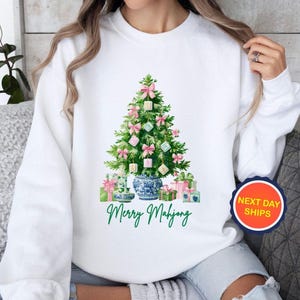 May include: White sweatshirt featuring a watercolor Christmas tree decorated with pink bows and square ornaments. The tree sits in a blue and white vase, with wrapped gifts at the base. The text "Merry Mahjong" is written in green.