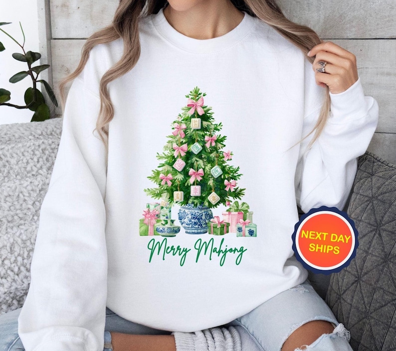 May include: White sweatshirt featuring a watercolor Christmas tree decorated with pink bows and square ornaments. The tree sits in a blue and white vase, with wrapped gifts at the base. The text "Merry Mahjong" is written in green.