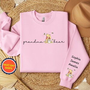 May include: A light pink sweatshirt with the words "grandma bear" in script. The design includes a cartoon bear with a honey pot and bees. The right sleeve has the names Sophia, James, and Amelia.