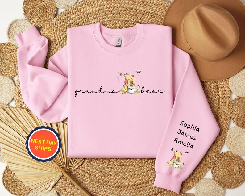 May include: A light pink sweatshirt with the words "grandma bear" in script. The design includes a cartoon bear with a honey pot and bees. The right sleeve has the names Sophia, James, and Amelia.