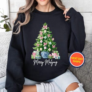 May include: Black sweatshirt featuring a Christmas tree design made of Mahjong tiles, pink bows, and wrapped gifts. The words "Merry Mahjong" are written in white script below the tree. A round sticker with the words "NEXT DAY SHIPS" is on the right.