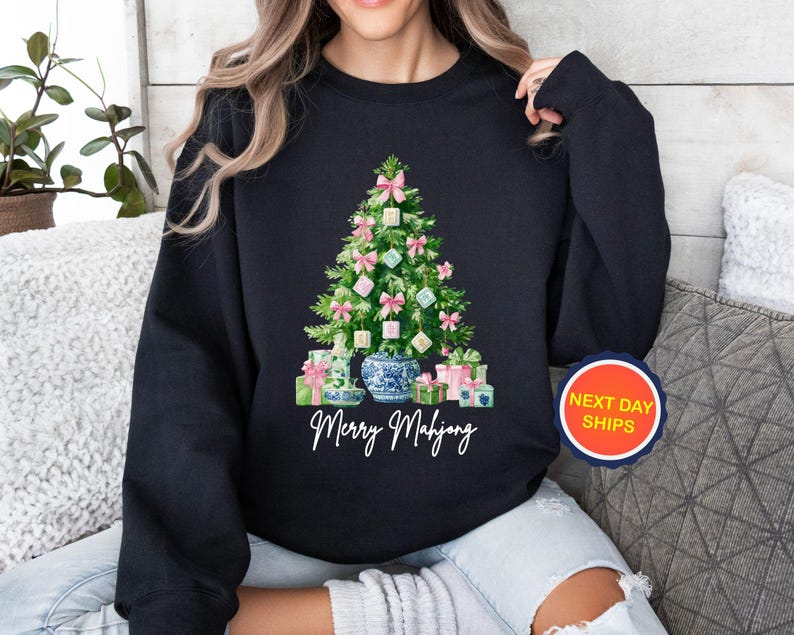 May include: Black sweatshirt featuring a Christmas tree design made of Mahjong tiles, pink bows, and wrapped gifts. The words "Merry Mahjong" are written in white script below the tree. A round sticker with the words "NEXT DAY SHIPS" is on the right.
