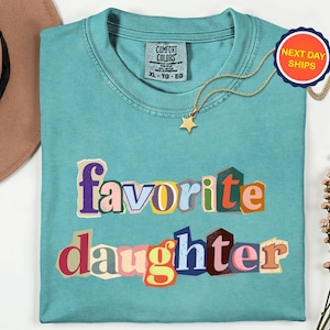 May include: A teal Comfort Colors t-shirt with the words "favorite daughter" in colourful, cut-out style letters. A gold star pendant necklace and a "Next Day Ships" sticker are also visible. The shirt is size XL-TG-EG.