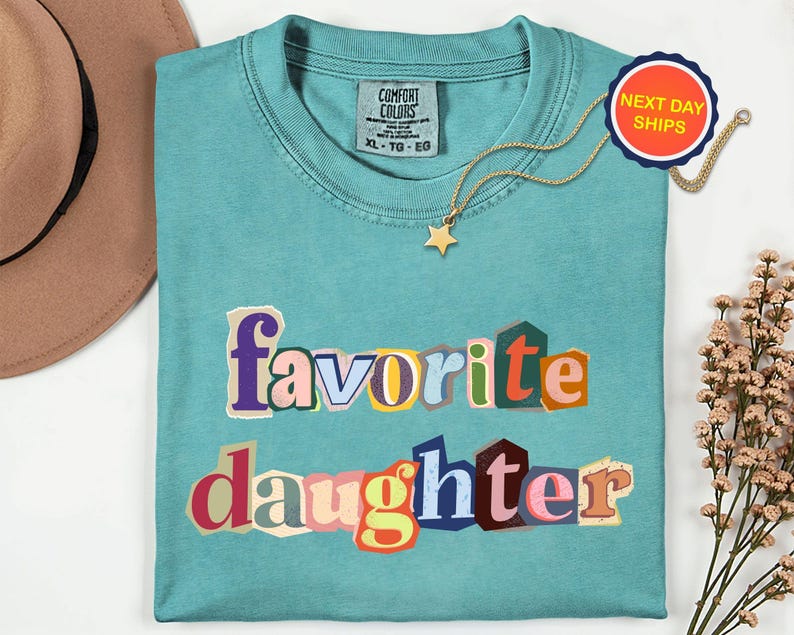 May include: A teal Comfort Colors t-shirt with the words "favorite daughter" in colourful, cut-out style letters. A gold star pendant necklace and a "Next Day Ships" sticker are also visible. The shirt is size XL-TG-EG.