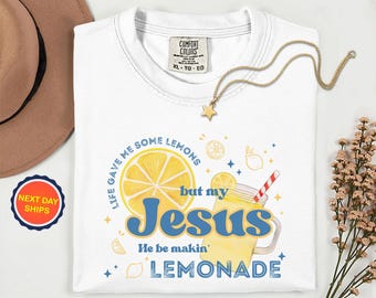 May include: White t-shirt with a graphic of a lemon slice and a lemonade drink. The text reads "Life gave me some lemons but my Jesus He be makin' LEMONADE". A gold star necklace is draped across the shirt.