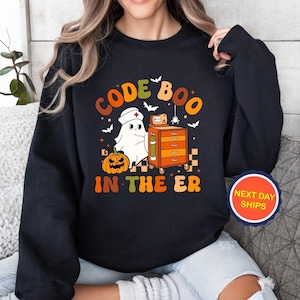 May include: Black sweatshirt with a Halloween-themed graphic. The design features a ghost wearing a nurse's cap, a pumpkin, and the text "CODE BOO IN THE ER" in orange and green. Bats and a spider are also included.