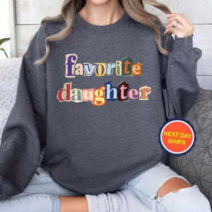 Puede incluir: Sudadera gris oscuro con las palabras "favorite daughter" en letras coloridas de estilo recortado. La sudadera tiene mangas largas y cuello redondo. Una pegatina circular con el texto "NEXT DAY SHIPS" es visible.