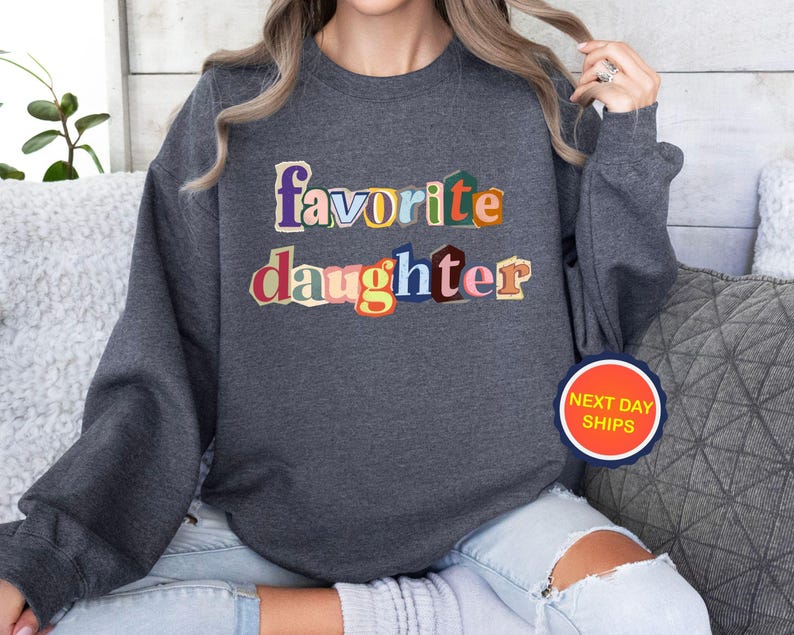 Puede incluir: Sudadera gris oscuro con las palabras "favorite daughter" en letras coloridas de estilo recortado. La sudadera tiene mangas largas y cuello redondo. Una pegatina circular con el texto "NEXT DAY SHIPS" es visible.