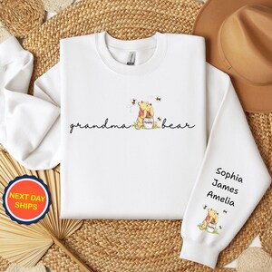 May include: White sweatshirt with the words "grandma bear" in cursive. The sweatshirt features a Winnie the Pooh graphic with bees and the names Sophia, James, and Amelia on the sleeve. A "Next Day Ships" sticker is visible.