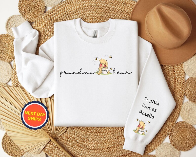 May include: White sweatshirt with the words "grandma bear" in cursive. The sweatshirt features a Winnie the Pooh graphic with bees and the names Sophia, James, and Amelia on the sleeve. A "Next Day Ships" sticker is visible.